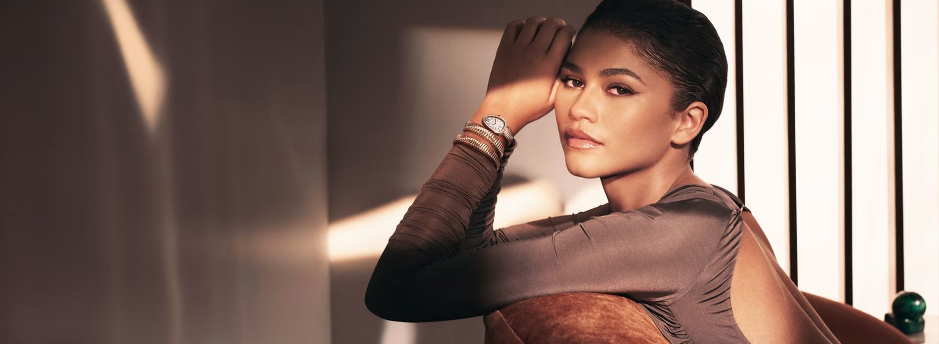 Zendaya leaning on a sofa and wearing a Serpenti Tubogas double-coil watch in yellow gold with a white dial.